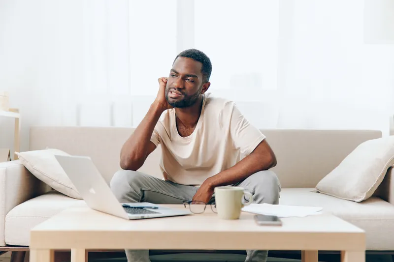 Уставший афроамериканский мужчина работает за ноутбуком в домашнем офисе