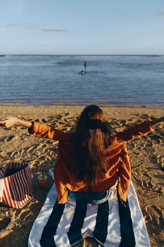 Woman on beach facing ocean horizon at sunset, warm sand light, arms open in relaxed pose, long dark hair, striped umbrella and striped towel, calm vacation mood
