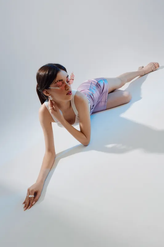 Young woman in stylish summer fashion lying on white floor wearing pink sunglasses and pastel outfit. Bright natural light highlights beauty and relaxed pose. Concept for fashion and beauty.