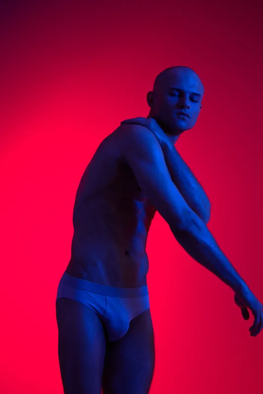 Athletic man posing confidently against a vibrant background, showcasing his physique with expressive body language, illuminated by contrasting lights.