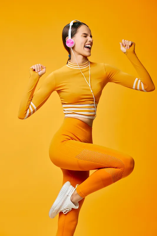A dynamic woman in bright orange activewear leaps midair in a studio, wearing headphones and a coordinated crop top and leggings, showcasing fitness, sport, movement, and energy during a bold workout