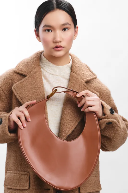 asian woman wears a cozy beige sweater and a brown coat, holding a curved leather bag with a calm, confident expression, posed against a white studio backdrop.
