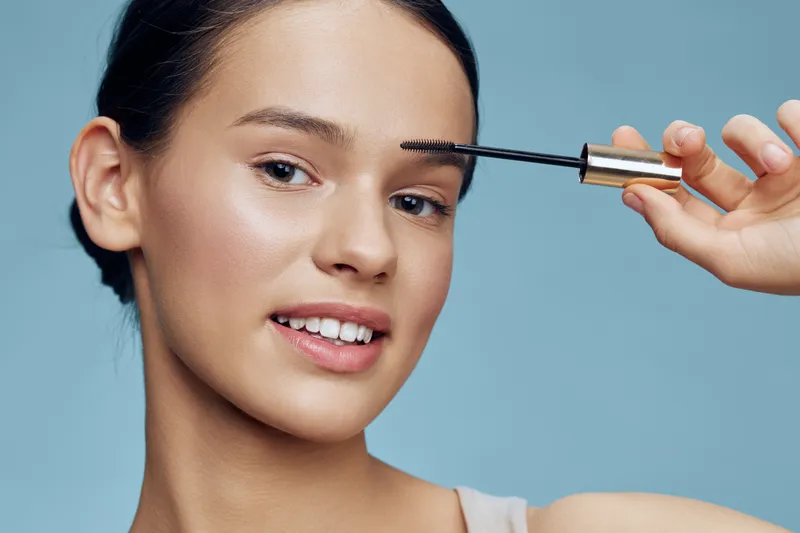 Young woman applying mascara, showcasing confidence and beauty against a minimalist blue background, emphasizing makeup and self care routines