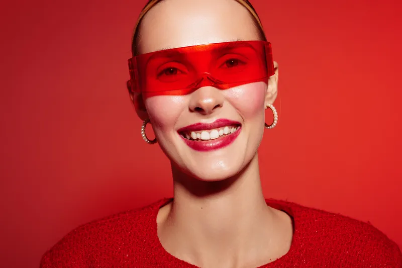 Smiling young woman wearing red sunglasses and red sweater poses against red background. Bright makeup and hoop earrings complete the vibrant fashion portrait concept.