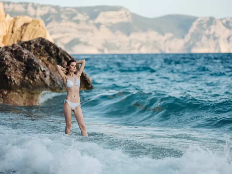 Красивая женщина в купальнике на пляже, рядом прозрачная вода океана и скалы, путешествия и туризм