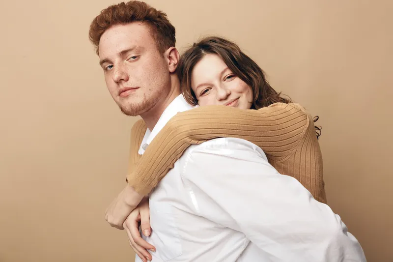 Young couple in casual clothes embraces against beige background. Woman hugs man from behind with smile, man looks confident. Concept of love, relationship, affection, and togetherness.