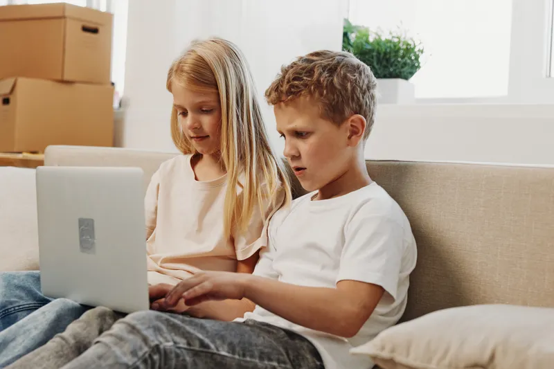 Two focused children, boy and girl, sitting on a sofa using a laptop together in a bright room with moving boxes in the background. Concept of learning, technology, and family bonding.