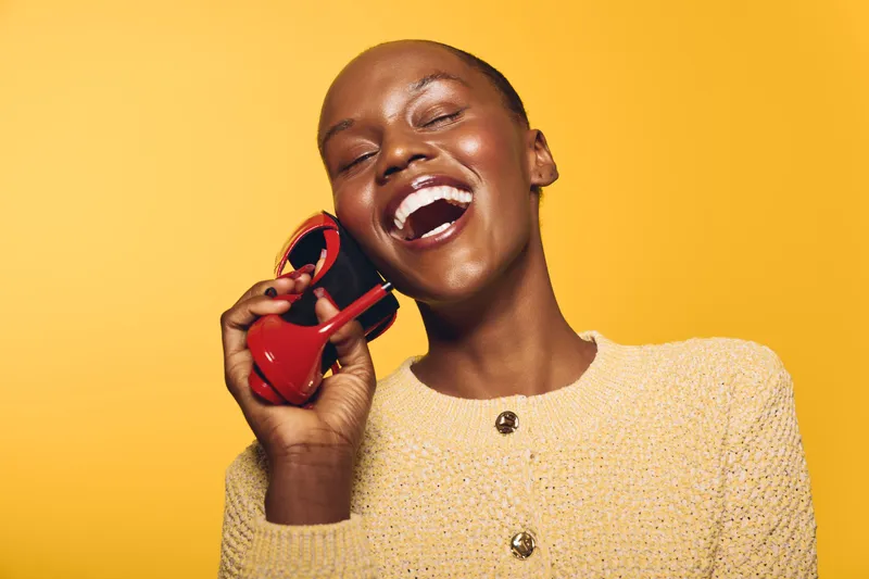 Joyful African woman with dark skin laughing and holding red sunglasses near her face. Studio portrait on yellow background. Concept of happiness, fashion, and summer style.