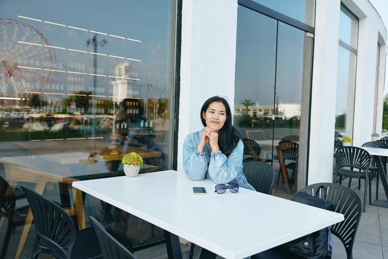 Outdoor cafe scene with a thoughtful woman seated at a white table, denim jacket, sunglasses, glass windows reflecting city life and calm atmosphere during bright afternoon moments.
