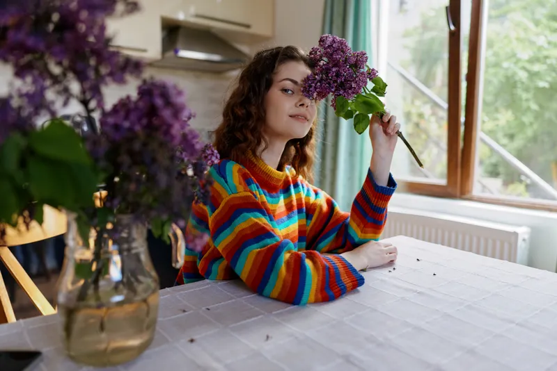 Young woman with curly hair sits at a bright home dining table, holding a colorful bouquet near a window and smiling softly.
