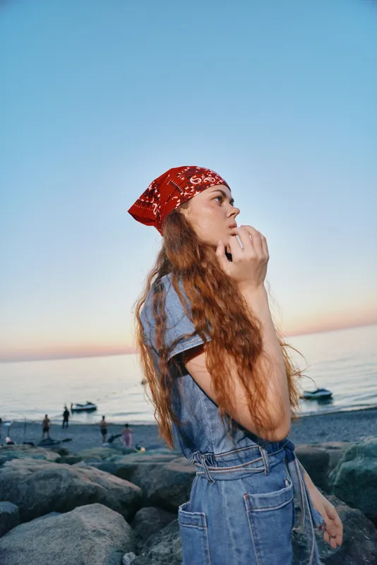 Lifestyle woman wearing modern boho-western denim street style outfit with red bandana, posing thoughtfully by the beach during sunset in film color tones.