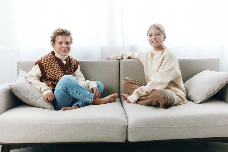 Two young children sit on a light gray sofa indoors, wearing cozy knit sweaters. A bright, airy room frames a calm scene of friendship and comfortable home life.