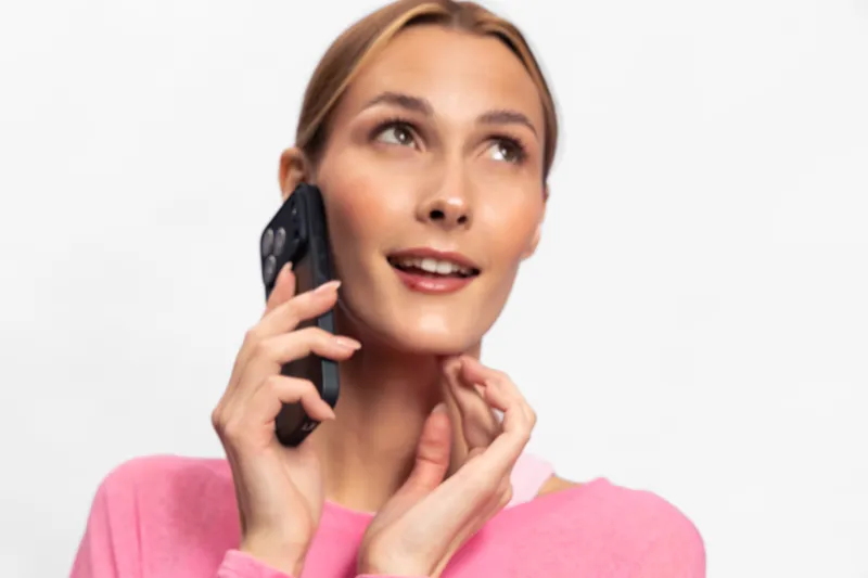 Young woman in pink sweater talking on smartphone, looking up with smile. Concept for communication, happiness, technology, and casual lifestyle with bright background.