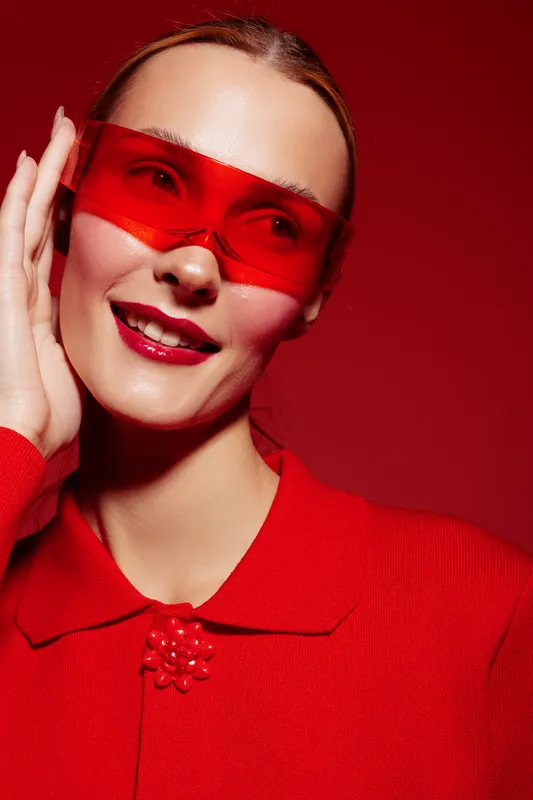 Smiling woman in red sunglasses and red outfit poses against red background. Fashion portrait with bright makeup and stylish accessories. Concept of modern style and confidence.
