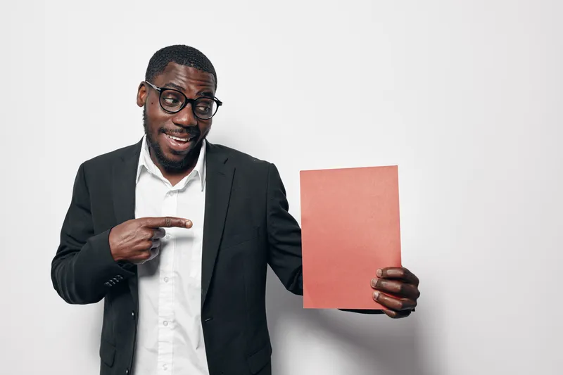 African American man with dark skin wearing glasses and black suit jacket holding and pointing at blank red paper sheet on white background for presentation or advertisement.