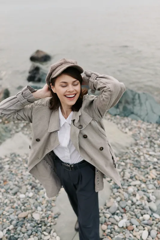 Happy young woman in casual autumn outfit smiles with closed eyes on rocky beach. She wears a patterned jacket and cap, enjoying nature and fresh air by the water shore.
