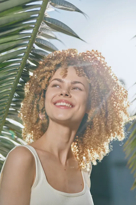 smiling woman with curly hair enjoys sunny outdoor atmosphere surrounded by green palm leaves, natural light highlights fresh and joyful mood