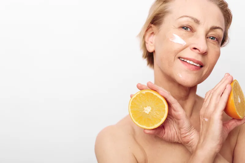 Mature woman with short hair holds two orange halves near face with cream on cheek. Concept of natural skincare, healthy lifestyle, and vitamin c beauty treatment.
