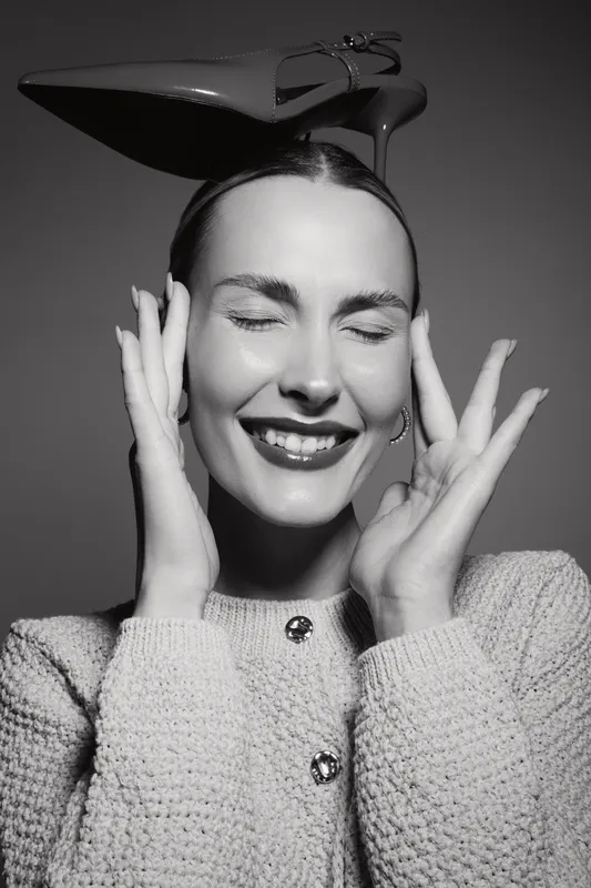 Happy white woman with fair skin smiling with closed eyes wearing knitted sweater and high heel shoe on head in studio. Concept of joy and fashion fun.