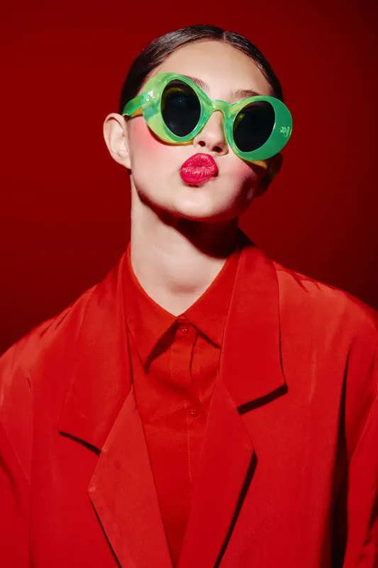Young woman with shiny red lipstick wearing oversized green sunglasses and red blazer posing on bright red backdrop. Concept of bold beauty creative fashion style.