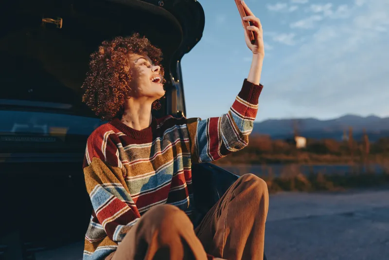 Woman lifestyle outdoors, roadtrip scene, curly hair by open car door, hand raised toward the sky, warm sunset light, mountains in distance, relaxed travel moment, wind and joy