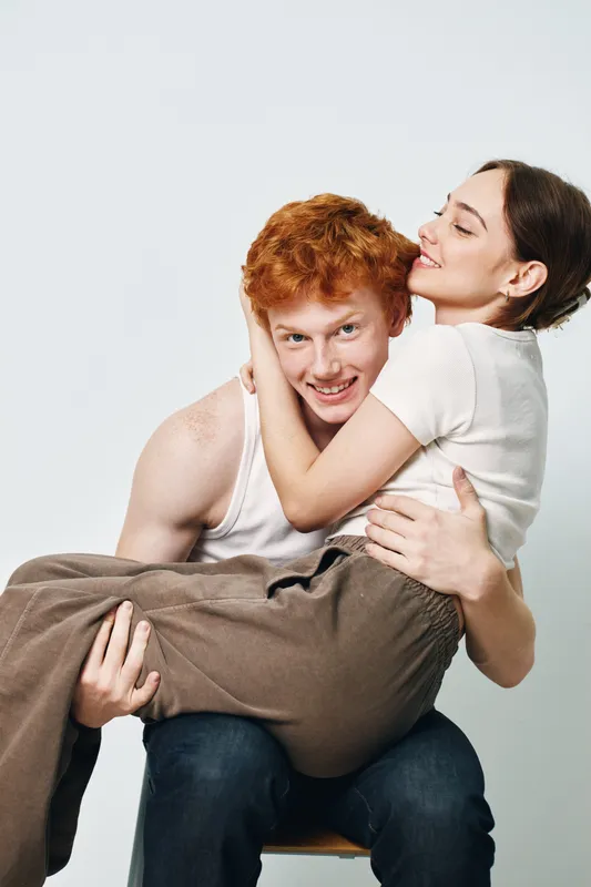 Young couple with red hair and brunette smiling and hugging, man holding woman in arms, lifestyle and affection with casual clothing in studio with white background.