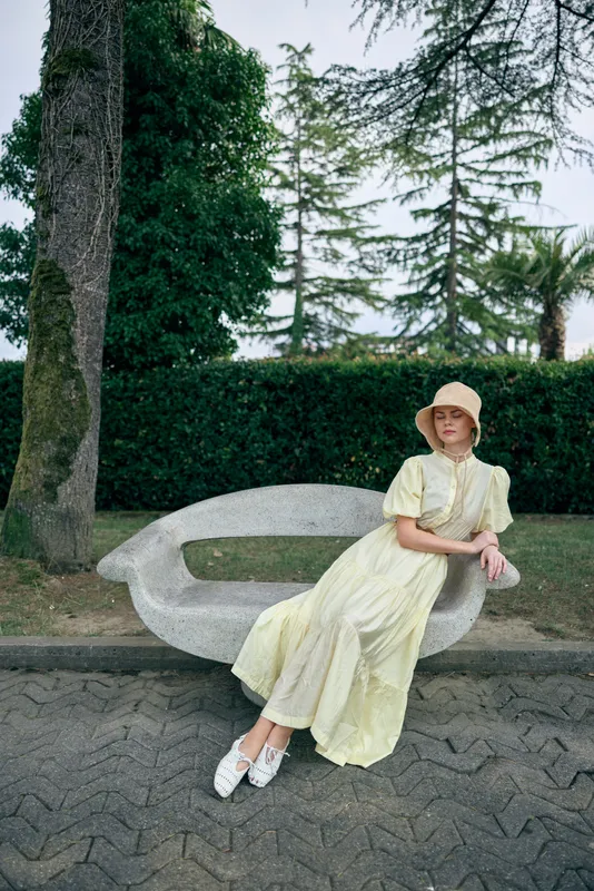 A woman sits on a park bench in a pale yellow dress and hat, enjoying a sunny day in a serene outdoor setting, exuding calm elegance and relaxed charm and poise.