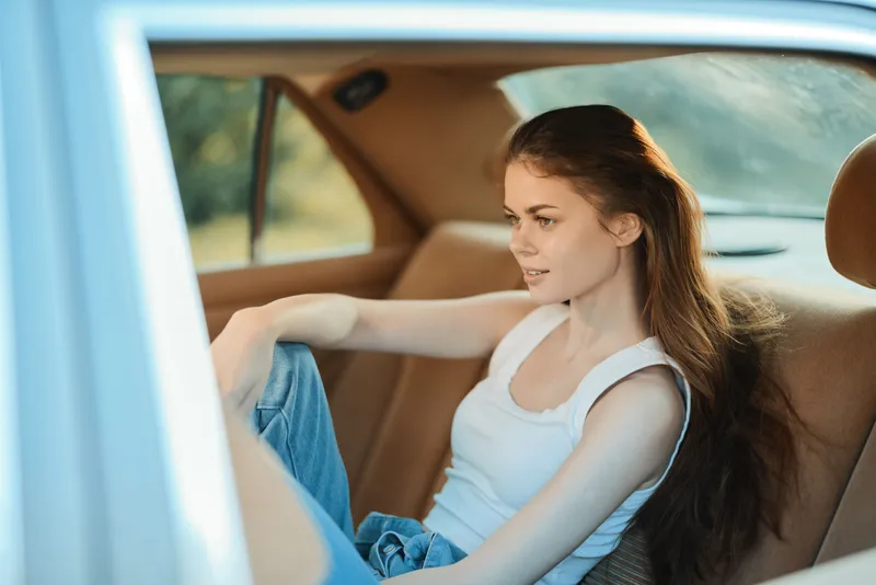 Young woman sitting in a car with relaxed expression, wearing a casual tank top and jeans, showcasing natural beauty and serenity in a sunny outdoor setting.