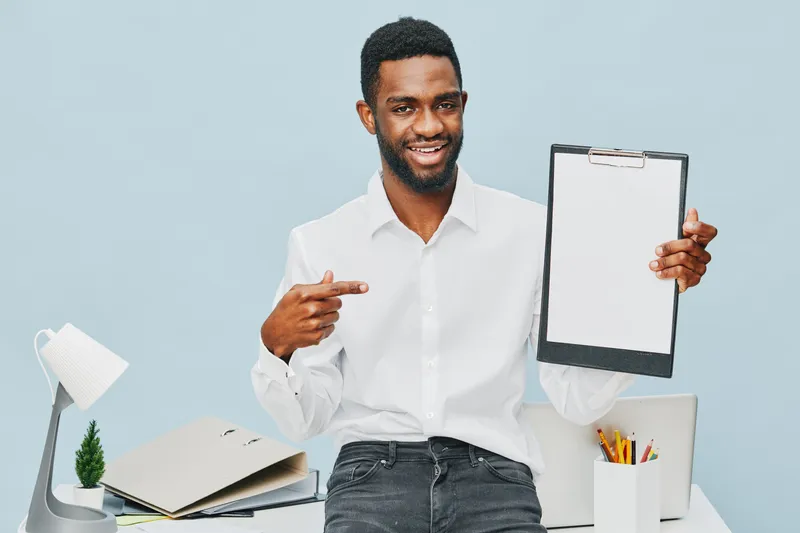 Smiling man with dark skin, wearing white shirt, pose with clipboard and pointing at it, seated at modern workspace with desk lamp, laptop, stationery, bright background. Business lifestyle concept.