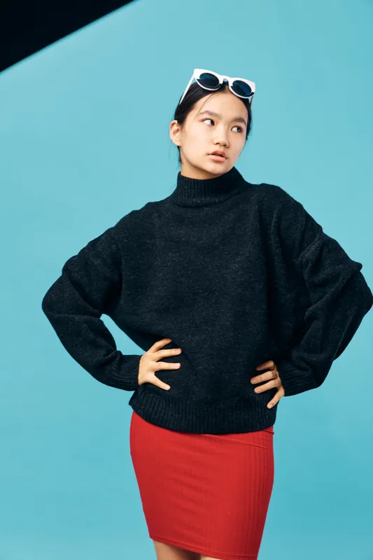 Young woman in black sweater and red skirt posing with hands on hips against blue background. Fashion portrait with casual style and thoughtful expression for modern look.