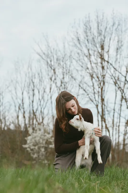 Young woman kneels in a grassy meadow, cuddling a small white dog. Soft natural light highlights companionship, happiness, and gentle affection in a tranquil setting.