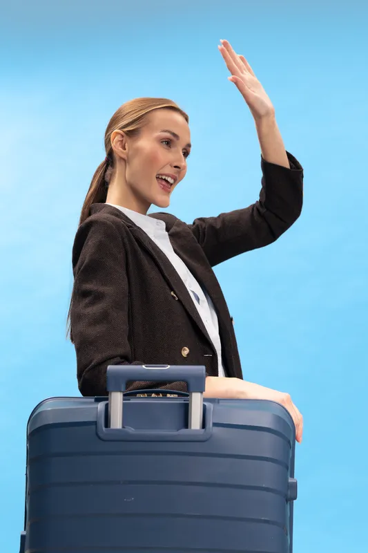 Young woman in black blazer waves hand smiling while standing with blue suitcase on blue background. Concept for travel, business trip, vacation and journey.