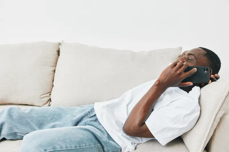 Young Black man relaxing on a couch while talking on a smartphone, dressed casually in a white t shirt and light jeans, exuding a calm and laid back emotion
