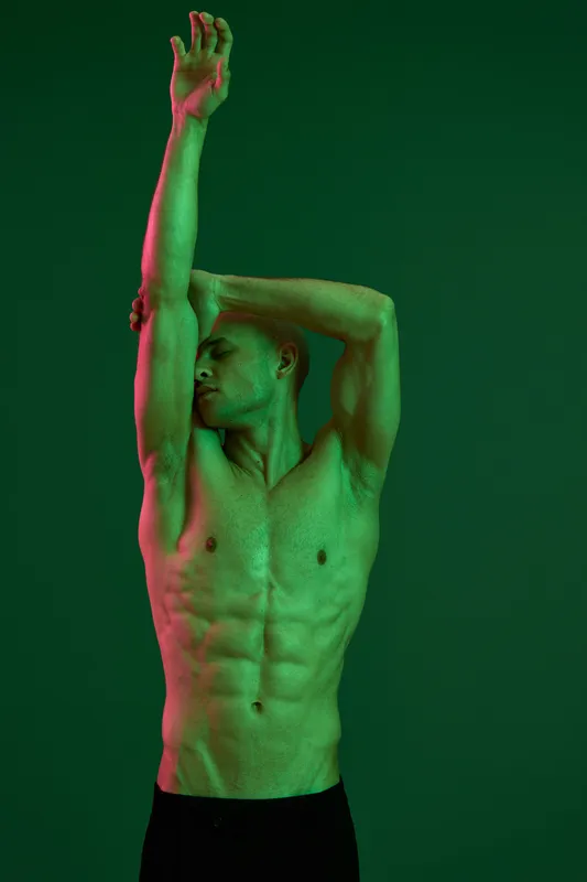Athletic man posing dramatically with green and pink lighting, highlighting strong muscles and body contours, showcasing dynamic form and movement in a vibrant setting.