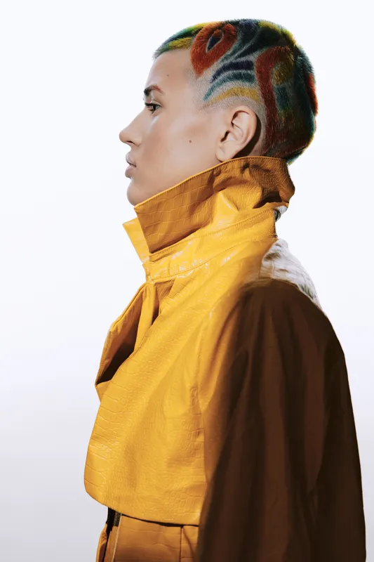 Woman with colorful shaved hair wearing a vibrant yellow jacket, featuring strong side profile, modern fashion style, and artistic expression in studio light.