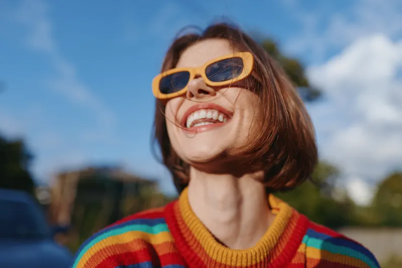 Smiling woman in yellow sunglasses and rainbow sweater, closeup portrait outdoors with bright sunlight, joyful expression and carefree fashion vibe for summer lifestyle shots.