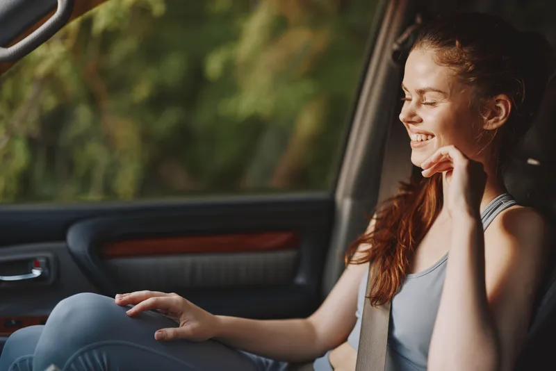 Young woman smiling in a car, showcasing casual fitness attire on a sunny day, representing joy and relaxation in a natural setting