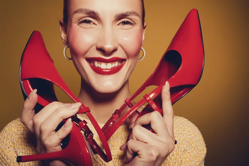 Smiling woman with red lipstick holds red high heel shoes close to her face on yellow background. Concept of fashion, style, beauty, and happiness with bright colors.