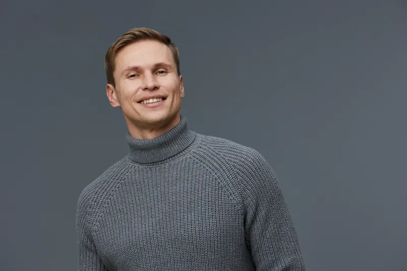 Smiling man with brown hair and blue eyes wearing a cozy gray turtleneck sweater on plain gray background. Casual, cheerful, relaxed, positive, fashion, lifestyle, portrait.