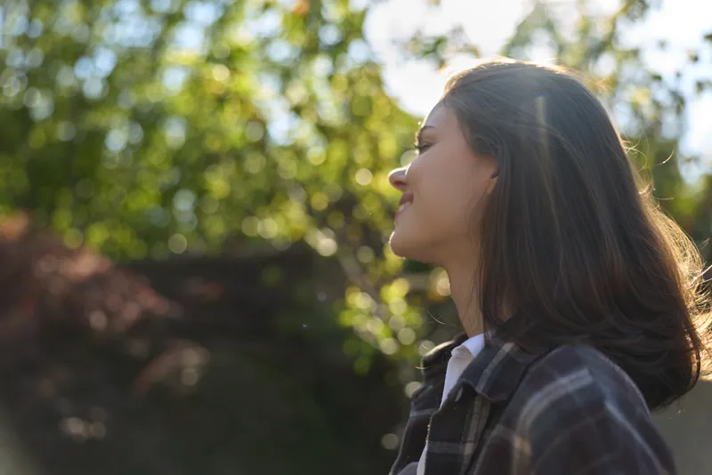 Happy woman outdoors, side profile smiling, sunlit moment, casual jacket, gentle breeze, natural tones, leafy background, candid lifestyle vibe