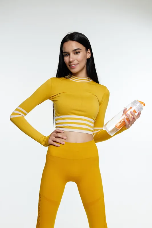 Woman sport fitness wearing yellow activewear holding water bottle, smiling confidently with long dark hair on clean white background, healthy lifestyle concept.