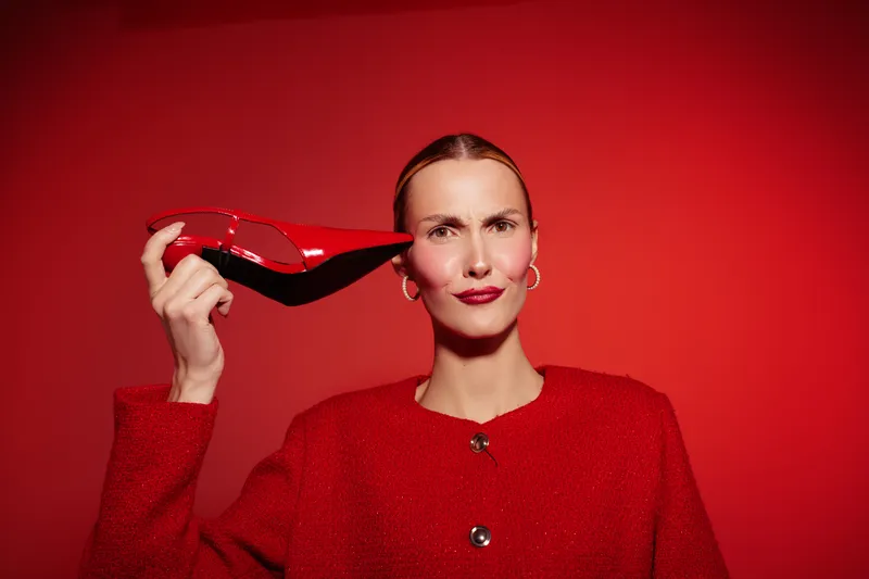 Woman in red coat holds red high heel shoe near ear on red background. Concept of fashion, style, and playful attitude with vibrant monochrome look.