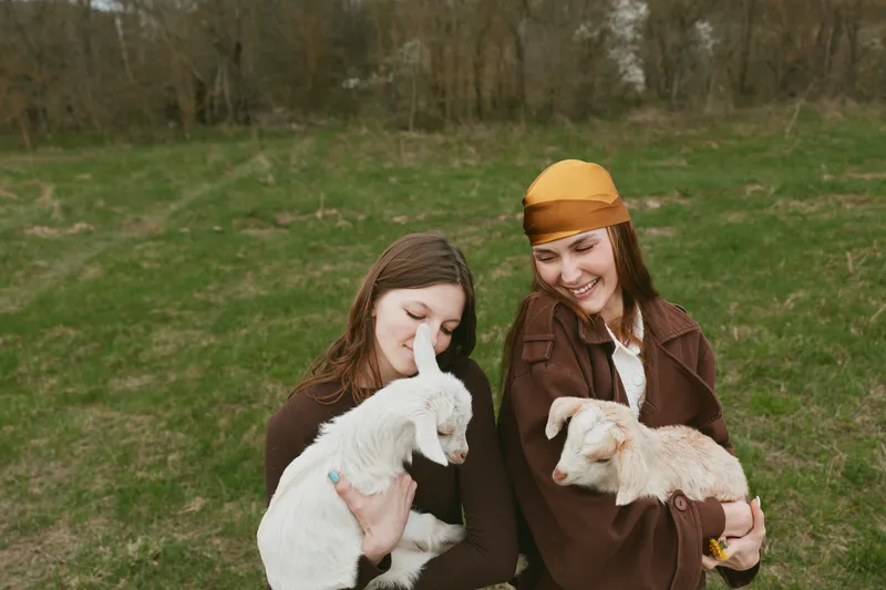 Two young women dressed in brown coats smile happily while holding white and light brown baby goats in a green meadow during a relaxed lifestyle moment outdoors.