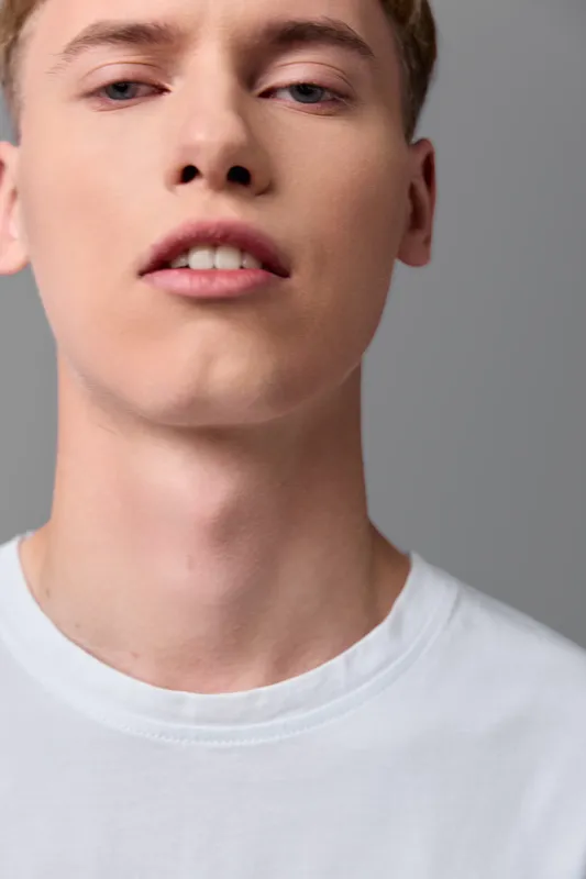 Close up of a young man in a white shirt with a neutral expression, studio lighting and soft gray background, highlighting facial features and skin texture in a clean minimalist frame