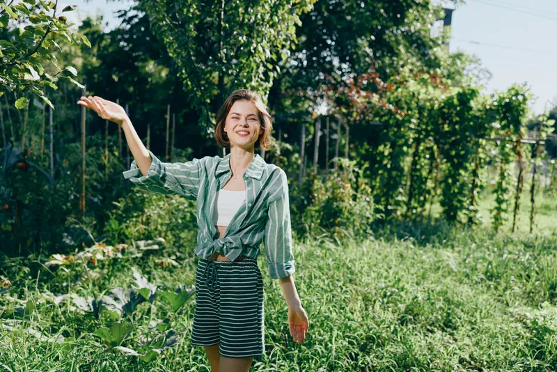 Young woman stands in a sunlit garden, smiling with arms outstretched, wearing a green shirt and striped shorts, surrounded by vibrant greenery and warm natural light.