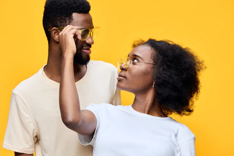 Happy multicultural couple wearing glasses on bright yellow background, showcasing love and connection through playful interaction and vibrant colors