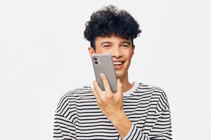 young man smiling holding smartphone, wearing striped shirt, curly hair, casual style, white background, modern technology use.