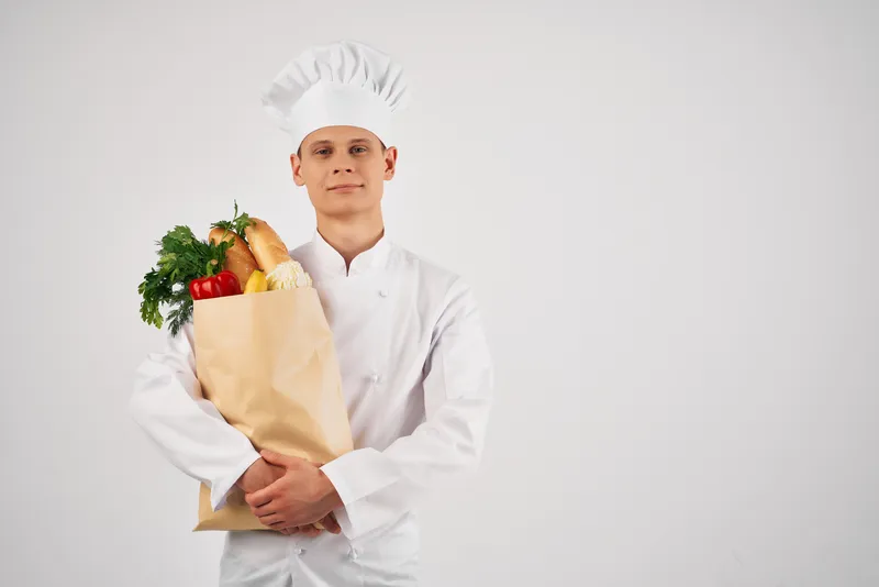 Весёлый мужчина-шеф-повар с пакетом продуктов здорового питания, предоставляющий услуги на кухне
