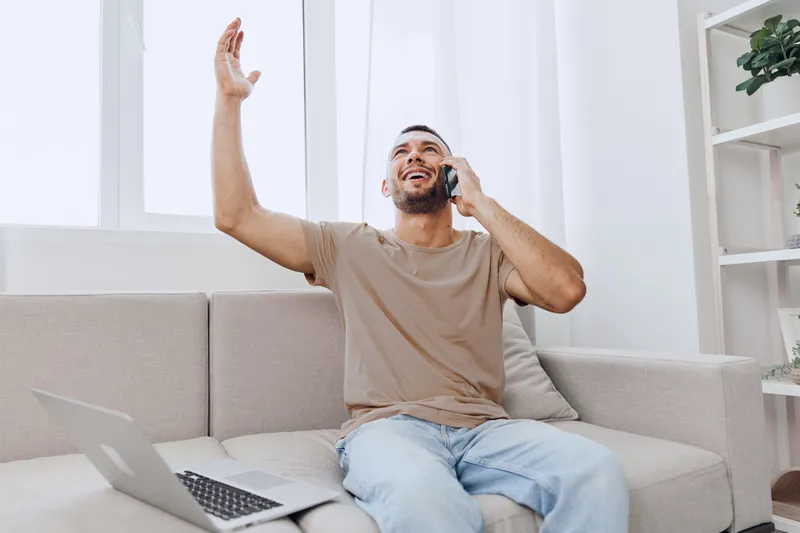 Excited man engaging in a phone conversation at home, with a laptop nearby and a bright, modern interior, conveying joy and productivity in a cozy environment.