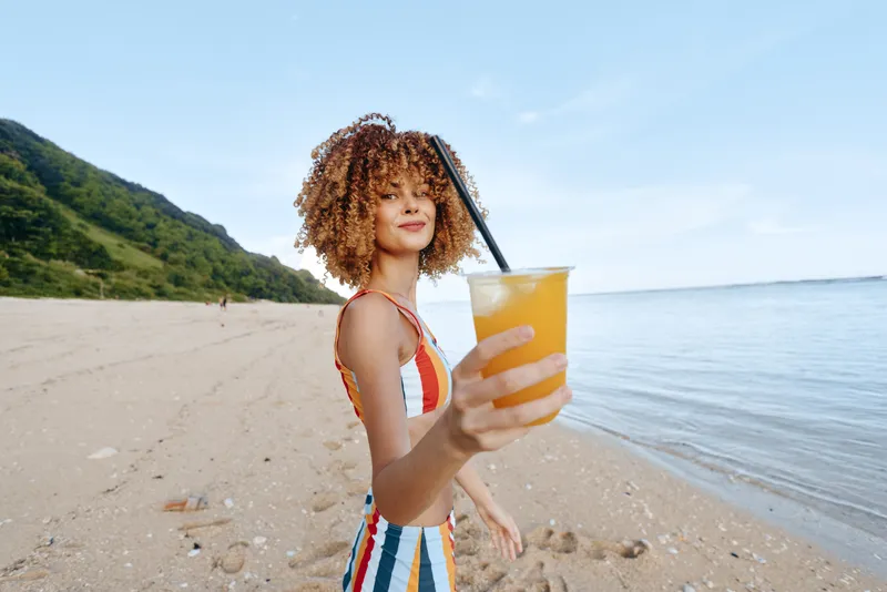 Woman with curly hair on a beach, holding orange juice in a glass. Summer seaside in bright sunlight, relaxed smile, outdoor refreshment and wellness.